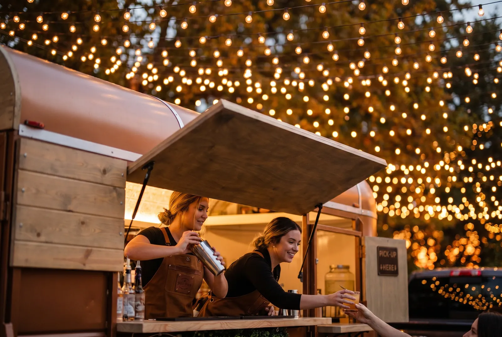 Two Steel Pony bartenders serving cocktails from the trailer under string lights at dusk