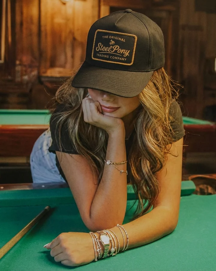 A woman leaning on a pool table wearing a black Steel Pony hat