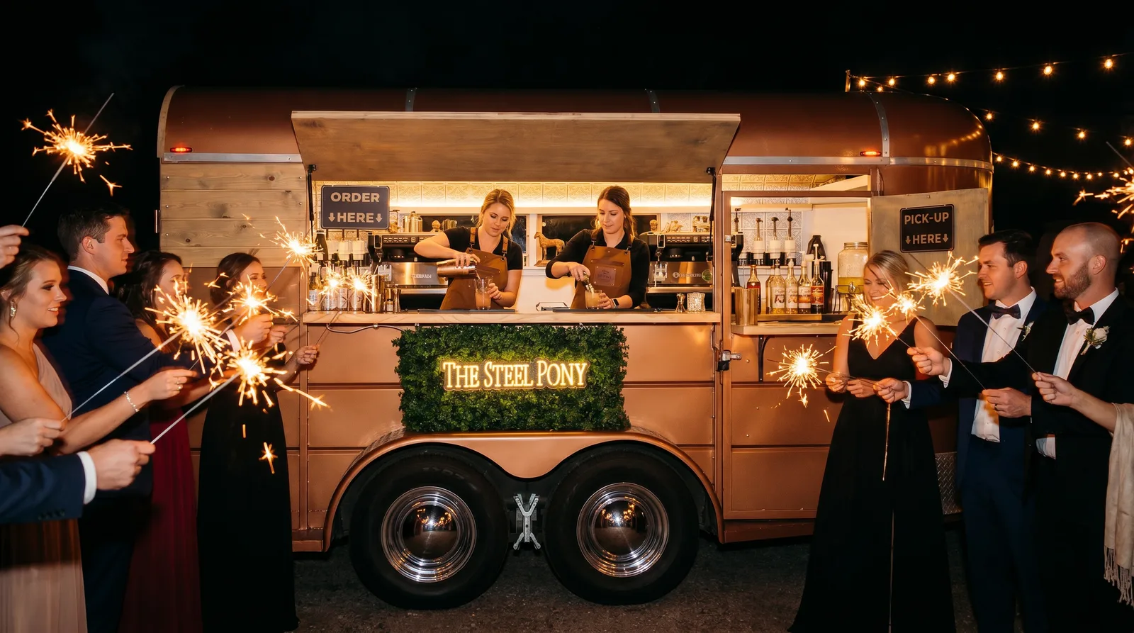 The wedding party holding sparklers around the Steel Pony horse-trailer bar at night