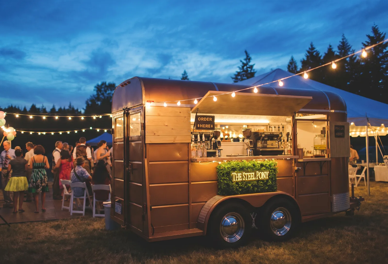 The trailer bar glowing at blue hour with the Order Here sign and guests nearby